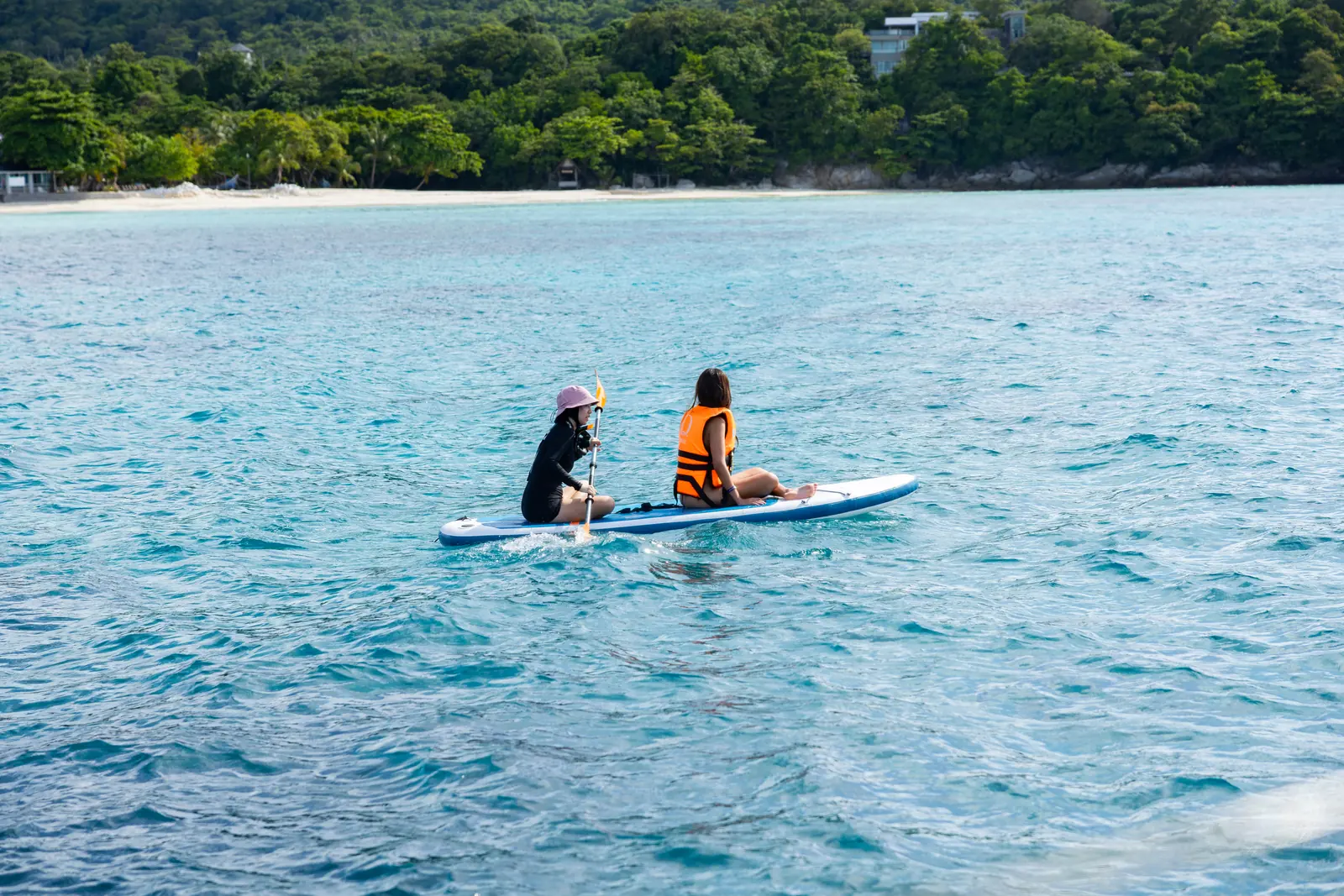 Two guests sharing a paddleboard on calm blue water near a wooded beach
