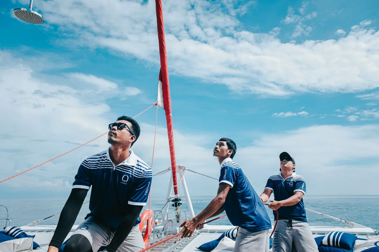 Three OMBRE crew members handling equipment on deck beneath a blue sky