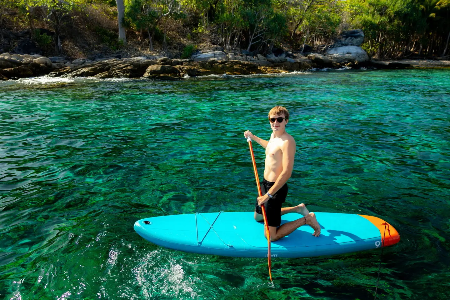 A guest kneeling on a turquoise paddleboard beside a rocky tree-lined shore